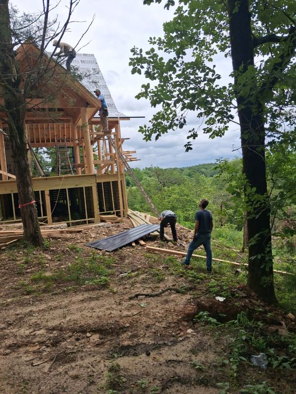 Crew building a timber frame cabin on a wooded hillside, with metal roof panels on the ground