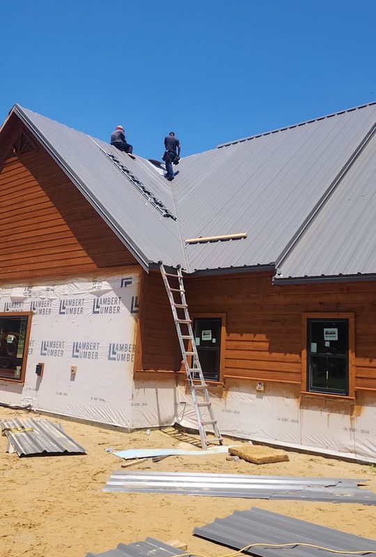 Two crew members installing a gray metal roof on a new house with cedar siding, a ladder against the wall and metal panels on the ground