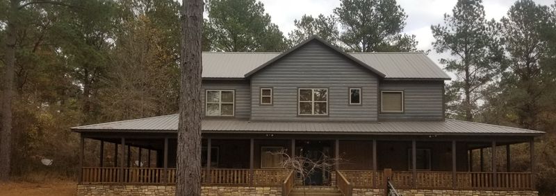 Large two story gray home with a metal roof and a wraparound porch, surrounded by pine trees